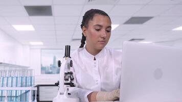 A young female researcher in a white coat looks at biological samples under a microscope and writes the results of a study to a laptop while sitting at a table in a science lab. video