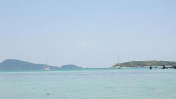 beautiful tropical sea under clear sunny blue sky with green island among turquoise sea with some boat, yacht in summer holiday vacation time. video