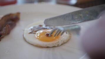 using knife and fork cutting egg yolk to eat fried egg in white plate. Cutting sunny side up egg with runny egg yolk video