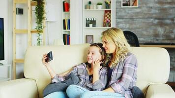 Little girl and her mother sitting on the couch in living room during a video call on the phone.