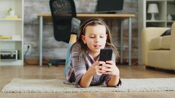 allegro poco ragazza dire bugie su il pavimento tappeto utilizzando smartphone. video