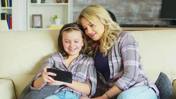 joven madre y su hija relajante en el sofá acecho un gracioso vídeo en teléfono inteligente video