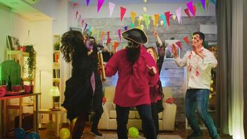 Bearded man dressed up like a pirate at halloween party blowing up confetti while his friends are having fun in the background. Slow motion shot video