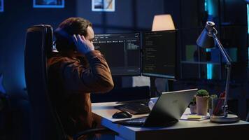 Technician focused on typing complex code in neon lit home office, developing software application. It specialist removing headphones to better concentrate on difficult task, camera A video