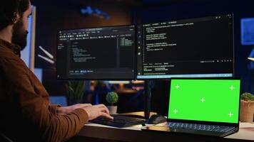 Programmer coding on computer next to isolated screen laptop in home office, developing software application. Teleworking IT specialist doing programming on PC near chroma key notebook, camera B video