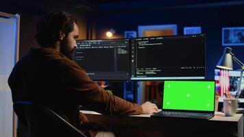 Green screen laptop next to freelancing man doing IT support job from apartment office, coding on PC. Mockup notebook on desk where software engineer writes lines of code on computer at home, camera B video