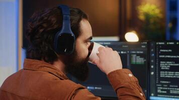 Man doing IT support job from apartment office, drinking cup of coffee and listening music while coding. Developer with headphones on enjoying hot tea and writing code on computer, camera A close up video