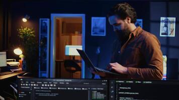 Man doing IT support job from home, walking around personal office with laptop in hand, coding. Nervous programmer wandering around room from stress, writing lines of code on notebook, camera B video