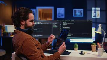 Software developer programming in home office, crosschecking lines of code between tablet and PC screen. IT admin holding portable device, using it as reference while coding on computer, camera B video