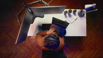 Top down view of engineer typing code in home office, using Java programming language to develop software application. It specialist using multimonitor PC setup and laptop to finish tasks, aerial shot video