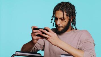 Man with disability in wheelchair playing videogames on cellphone, enjoying leisure time. middle eastern gamer enjoying game on mobile phone, having fun, studio background, camera B video