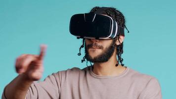 middle eastern man wearing virtual reality headset, doing swiping gestures. BIPOC person using high tech futuristic modern VR gadget, isolated over blue studio background, camera B video