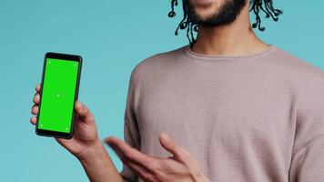 Happy man pointing towards green screen mobile phone, showing thumbs up sign. Cheerful person presenting chroma key cellphone, doing gesturing, isolated over studio background, camera B close up video