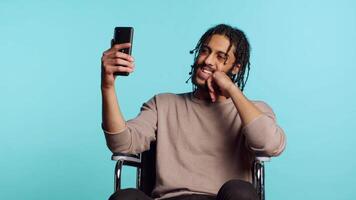 Man in wheelchair saluting friend over online videocall on cellphone, enjoying leisure time. Smiling person with disability bonding with mate using phone videoconference, studio background, camera A video