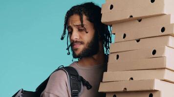 Portrait of smiling pizza delivery man holding takeaway orders for customer. Upbeat BIPOC courier with lunch boxes in arms and thermal food backpack, isolated over blue studio background, camera B video