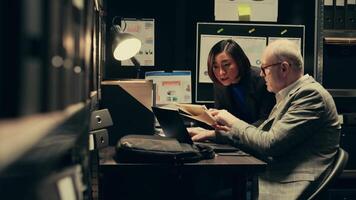 Law agents studying old classified information from police archives folders, looking for clues and crime trends. Partners accessing additional archived resources in incident room. Camera B. video