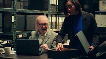 Senior inspector showing case details to his partner in archive room, studying forensic evidence and classified criminal records in a box. Detectives team work on federal investigation. Camera A. video