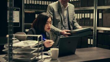 Senior inspector showing case details to his partner in archive room, studying forensic evidence and classified criminal records in a box. Detectives team work on federal investigation. Camera B. video