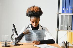 hembra africano americano médico vistiendo blanco Saco con estetoscopio sentado detrás escritorio en el oficina y mirando radiografía imagen. foto