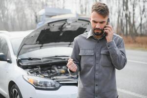 hombre utilizar un Teléfono móvil llamada garaje en frente de el abierto capucha de un roto coche en el la carretera en el bosque. coche Descompostura concepto. foto