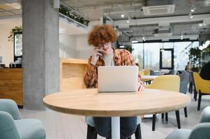 Young happy male freelancer in casual clothes sitting in cafe with laptop and using mobile phone. photo