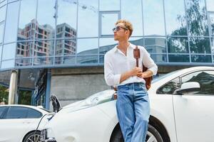 hombre cargando su lujo eléctrico coche a al aire libre estación en frente de moderno nuevo ciudad edificios foto