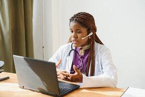 sonriente africano americano hembra médico gp usa blanco médico Saco utilizando ordenador portátil computadora a lugar de trabajo da remoto en línea consulta, trabajando en ordenador personal, consultante paciente en Internet telemedicina charla foto
