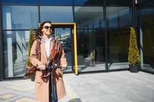 Young beautiful woman in a jacket smiles and rides an electric scooter to work along office buildings photo