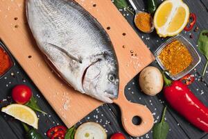 Raw sea fish dorado with lemon, pepper, tomatoes and various spices. On a black rustic background. photo