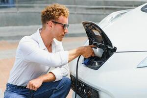 Hansome guy sitting near his new modern electric car and holding plug of the charger, while car is charging at the charging station photo