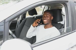 Happy smiling African American male driver sitting behind the self-driving steering wheel of an autonomous electric modern car. Happy guy holds phone and smiles to camera in modern electric car photo