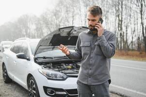 Sad driver calling car service, opening hood, having engine problem standing near broken car on the road. Car breakdown concept photo