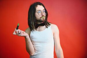 joven hombre aplicando labio brillo en su labios y mirando en cámara me gusta espejo. gay hombre haciendo hacer arriba en contra rojo antecedentes foto