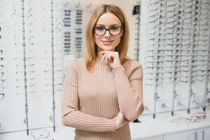 eyesight and vision concept - young business woman choosing glasses at optics store. photo