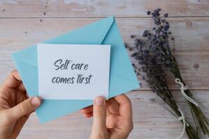 Female hands taking paper card note with text SELF CARE COMES FIRST from blue envelope. Lavender flower. Top view, flat lay. Concept of mental spiritual health self care wellbeing mindfulness photo