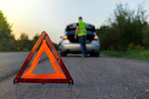 Broken silver luxury car emergency accident. Man driver installing red triangle stop sign on road. Sport automobile turned on blinkers technical problems on the road. Safety procedure when having vehicle broken down photo