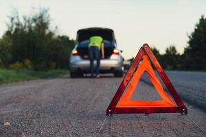 Unrecognizable sad driver in reflective vest. Male driver standing near broken car with open up hood. Red triangle to warn other road users of car breakdown or engine failure stop at countryside highway. Emergency traffic situation photo