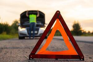 Unrecognizable sad driver in reflective vest. Male driver standing near broken car with open up hood. Red triangle to warn other road users of car breakdown or engine failure stop at countryside highway. Emergency traffic situation photo