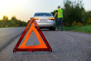 Unrecognizable sad driver in reflective vest. Male driver standing near broken car with open up hood. Red triangle to warn other road users of car breakdown or engine failure stop at countryside highway. Emergency traffic situation photo