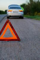 irreconocible triste conductor en reflexivo chaleco. masculino conductor en pie cerca roto coche con abierto arriba capucha. rojo triángulo a advertir otro la carretera los usuarios de coche Descompostura o motor fracaso detener a campo carretera. emergencia tráfico situación foto