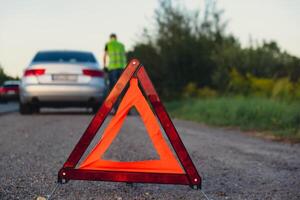 Broken silver luxury car emergency accident. Man driver installing red triangle stop sign on road. Sport automobile turned on blinkers technical problems on the road. Safety procedure when having vehicle broken down photo