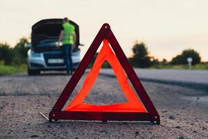 irreconocible triste conductor en reflexivo chaleco. masculino conductor en pie cerca roto coche con abierto arriba capucha. rojo triángulo a advertir otro la carretera los usuarios de coche Descompostura o motor fracaso detener a campo carretera. emergencia tráfico situación foto