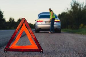 Unrecognizable sad driver in reflective vest. Male driver standing near broken car with open up hood. Red triangle to warn other road users of car breakdown or engine failure stop at countryside highway. Emergency traffic situation photo