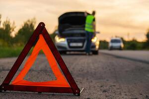 Unrecognizable sad driver in reflective vest. Male driver standing near broken car with open up hood. Red triangle to warn other road users of car breakdown or engine failure stop at countryside highway. Emergency traffic situation photo