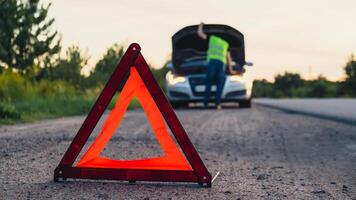 Broken silver luxury car emergency accident. Man driver installing red triangle stop sign on road. Sport automobile turned on blinkers technical problems on the road. Safety procedure when having vehicle broken down photo