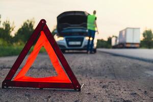 irreconocible triste conductor en reflexivo chaleco. masculino conductor en pie cerca roto coche con abierto arriba capucha. rojo triángulo a advertir otro la carretera los usuarios de coche Descompostura o motor fracaso detener a campo carretera. emergencia tráfico situación foto