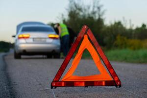 Unrecognizable sad driver in reflective vest. Male driver standing near broken car with open up hood. Red triangle to warn other road users of car breakdown or engine failure stop at countryside highway. Emergency traffic situation photo