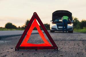 Unrecognizable sad driver in reflective vest. Male driver standing near broken car with open up hood. Red triangle to warn other road users of car breakdown or engine failure stop at countryside highway. Emergency traffic situation photo