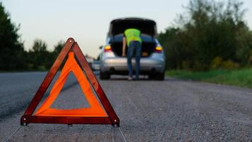 Broken silver luxury car emergency accident. Man driver installing red triangle stop sign on road. Sport automobile turned on blinkers technical problems on the road. Safety procedure when having vehicle broken down photo