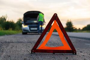 Broken silver luxury car emergency accident. Man driver installing red triangle stop sign on road. Sport automobile turned on blinkers technical problems on the road. Safety procedure when having vehicle broken down photo